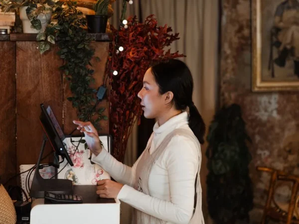 Side view of focused young ethnic female cashier wearing apron using modern cash desk with computer screen in cafeteria with rustic wooden interior in dark light in evening