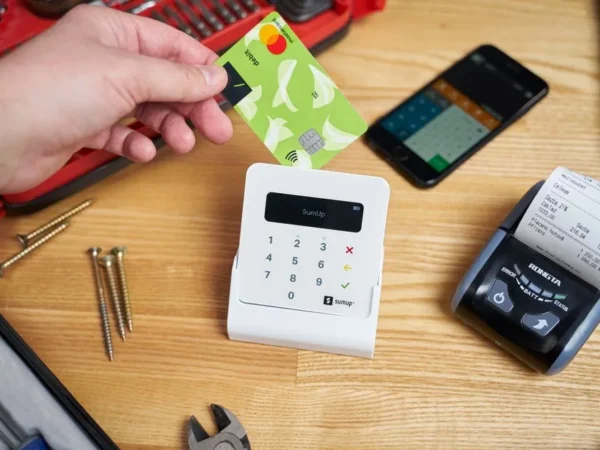 Faceless customer paying by small bright plastic card while using cash register among smartphone and tools on wooden table in bright studio