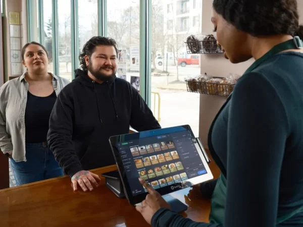 Barista using POS system to serve two customers at a café counter, showcasing modern restaurant technology.
